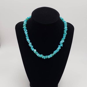 May include: A turquoise chip necklace displayed on a black mannequin. The necklace is composed of small, irregular-shaped turquoise stones, strung together to form a choker-style necklace. The stones are a vibrant shade of blue-green.