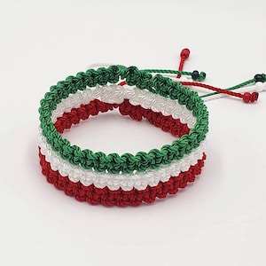 May include: A green, white, and red braided friendship bracelet. The bracelet is made of three strands of cord, each a different color. The cords are braided together to create a pattern.