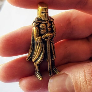 May include: A gold-colored metal figurine of a knight wearing armor and a cape. The knight is holding a sword in his right hand.