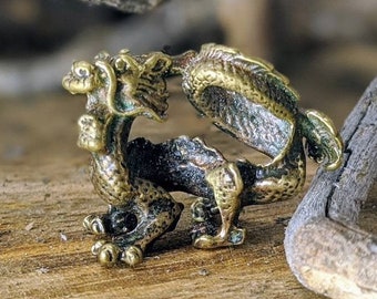 May include: A detailed, bronze-colored dragon ring. The ring features a dragon with intricate scales and details, posed in a circular design. The ring is set against a natural wood background with dried branches.