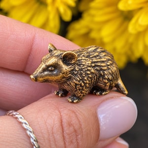 May include: A small, antique-style brass figurine of an opossum. The miniature animal sculpture features a textured, metallic gold-toned finish with dark highlights, showing detailed fur, pointed ears, a long snout, and a short tail.