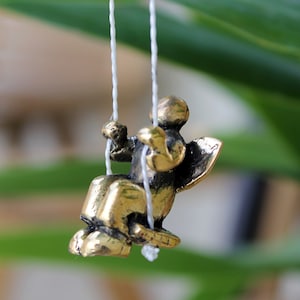 May include: A small gold-coloured figurine of an angel on a swing. The angel has wings and is holding onto the ropes of the swing. The swing is suspended from white strings.