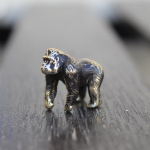 K&ouml;nnte beinhalten: Eine kleine, detaillierte Bronzefigur eines Gorillas, die auf einer dunklen Holzoberfl&auml;che steht.