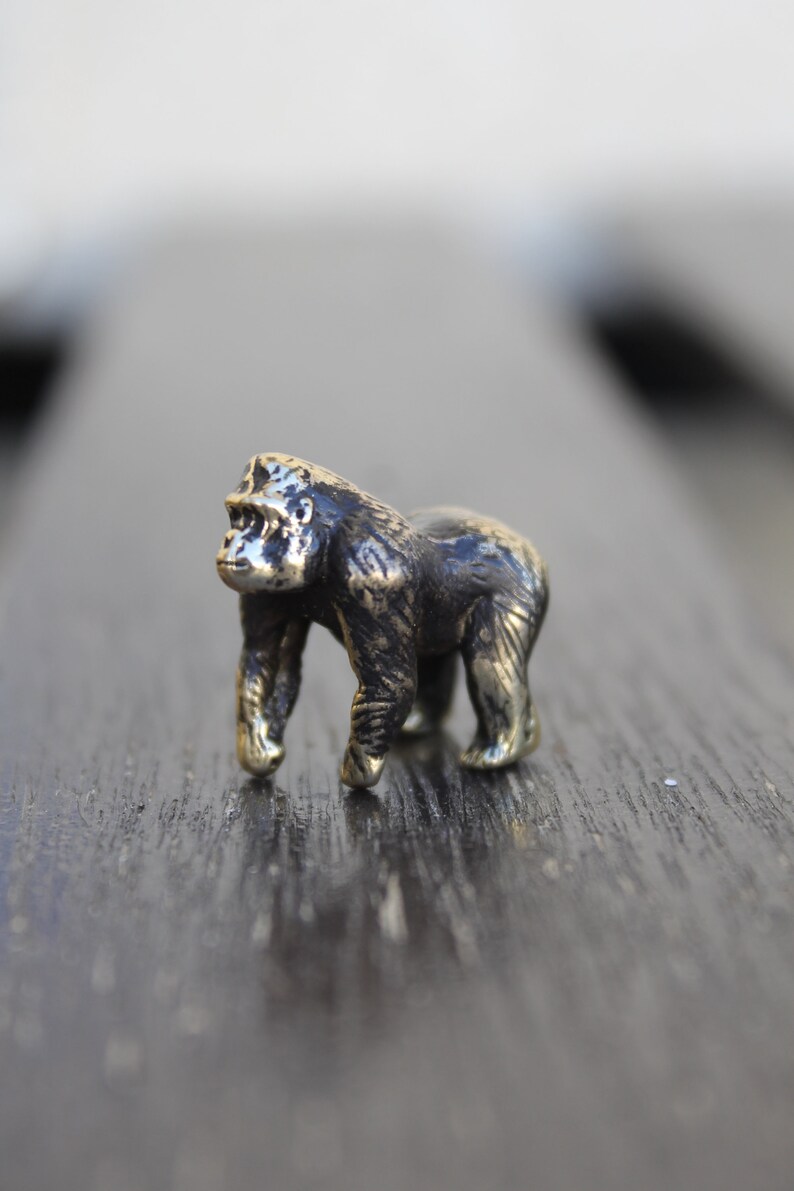 K&ouml;nnte beinhalten: Eine kleine, detaillierte Bronzefigur eines Gorillas, die auf einer dunklen Holzoberfl&auml;che steht.