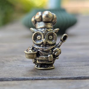 May include: A small, bronze figurine of an owl wearing a chef's hat and holding a spoon and a pot. The owl has large, round eyes and a detailed, textured surface.