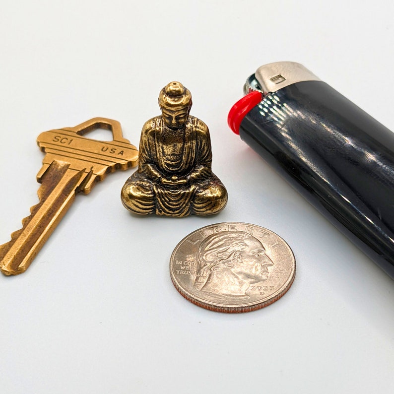 May include: A gold-colored metal Buddha statue sitting on a white surface. The statue is in a seated meditation pose with hands in the lap. There is a key, a lighter, and a US quarter coin in the image.