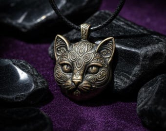 Pode incluir: Um pingente em forma de cabeça de gato em tom de bronze com padrões intrincados de redemoinhos e motivos oculares na testa e nas bochechas. O pingente está suspenso por um cordão preto simples, exibindo um acabamento metálico detalhado.