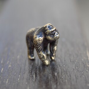 K&ouml;nnte beinhalten: Eine kleine, bronzefarbene Gorilla-Figur. Die Figur steht auf einer dunklen Holzoberfl&auml;che.