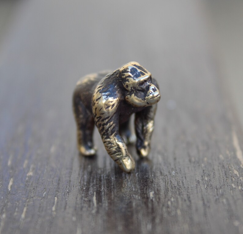 K&ouml;nnte beinhalten: Eine kleine, bronzefarbene Gorilla-Figur. Die Figur steht auf einer dunklen Holzoberfl&auml;che.