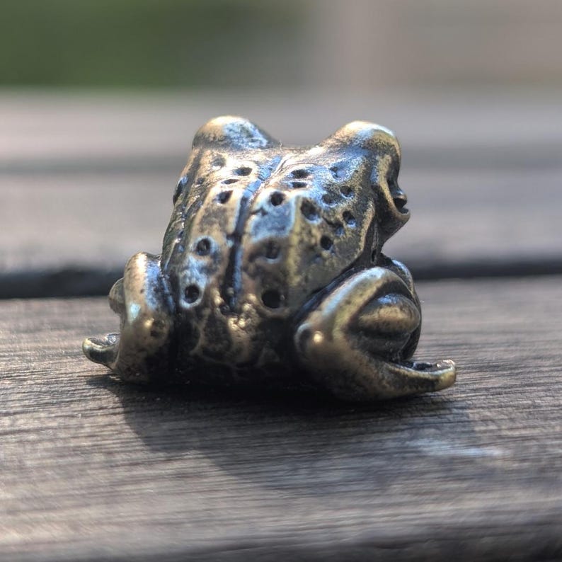 Pode incluir: Pequena e detalhada estatueta de r&atilde; em bronze. A r&atilde; est&aacute; sentada com pele texturizada e detalhes vis&iacute;veis dos olhos. O bronze tem uma apar&ecirc;ncia ligeiramente envelhecida, dando-lhe um toque vintage. &Eacute; um item decorativo.