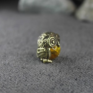 May include: A gold-colored owl-shaped earring with intricate details. The earring is made of metal and has a small, round, yellow stone in the center.