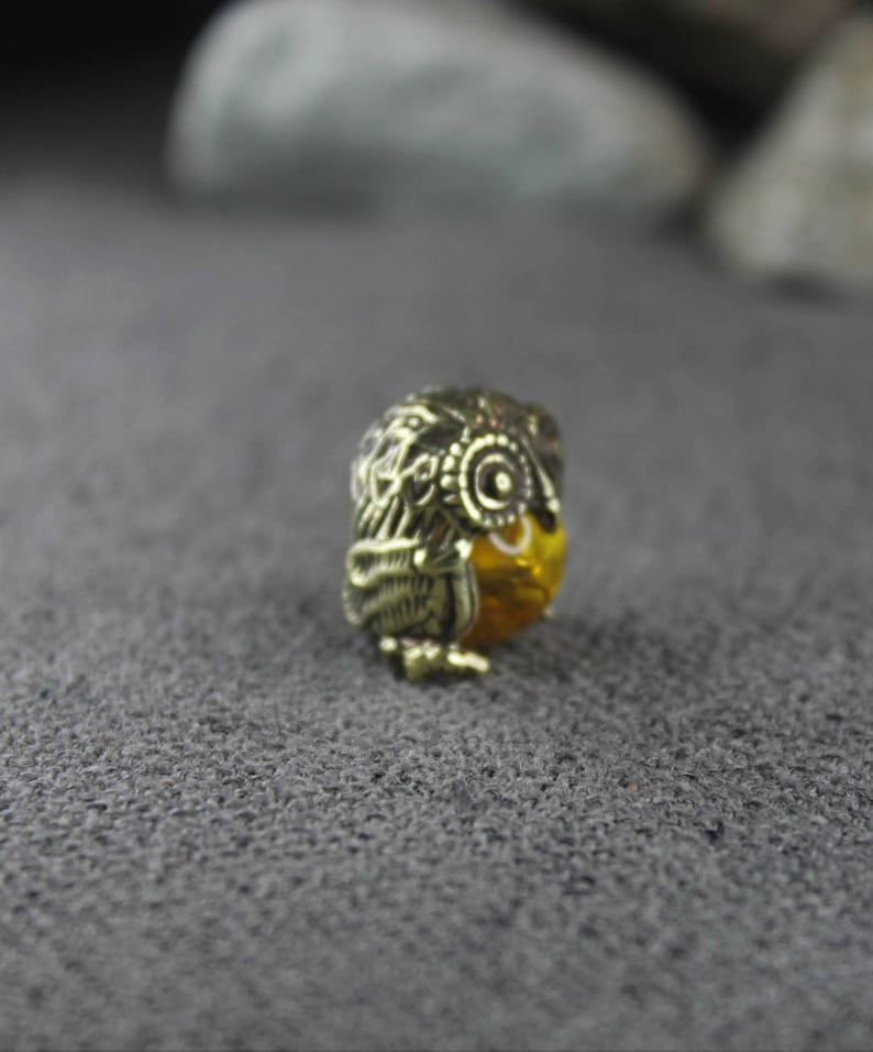 May include: A gold-colored owl-shaped earring with intricate details. The earring is made of metal and has a small, round, yellow stone in the center.
