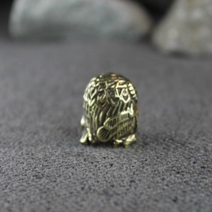 May include: A small, gold-colored metal thimble with an intricate design of an owl. The thimble is sitting on a gray, textured surface.