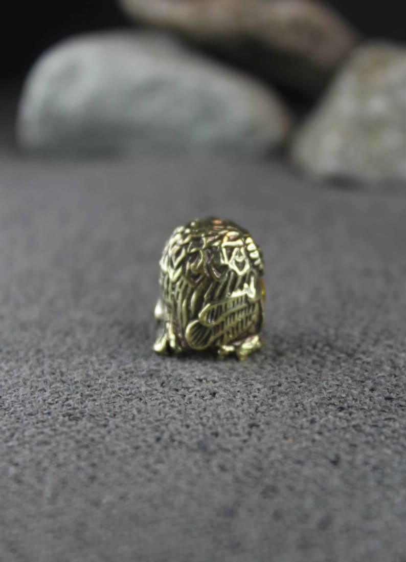 May include: A small, gold-colored metal thimble with an intricate design of an owl. The thimble is sitting on a gray, textured surface.