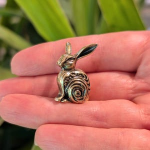 May include: A small, gold-colored metal rabbit figurine with intricate swirling patterns on its body. The rabbit is sitting upright with its ears perked up.