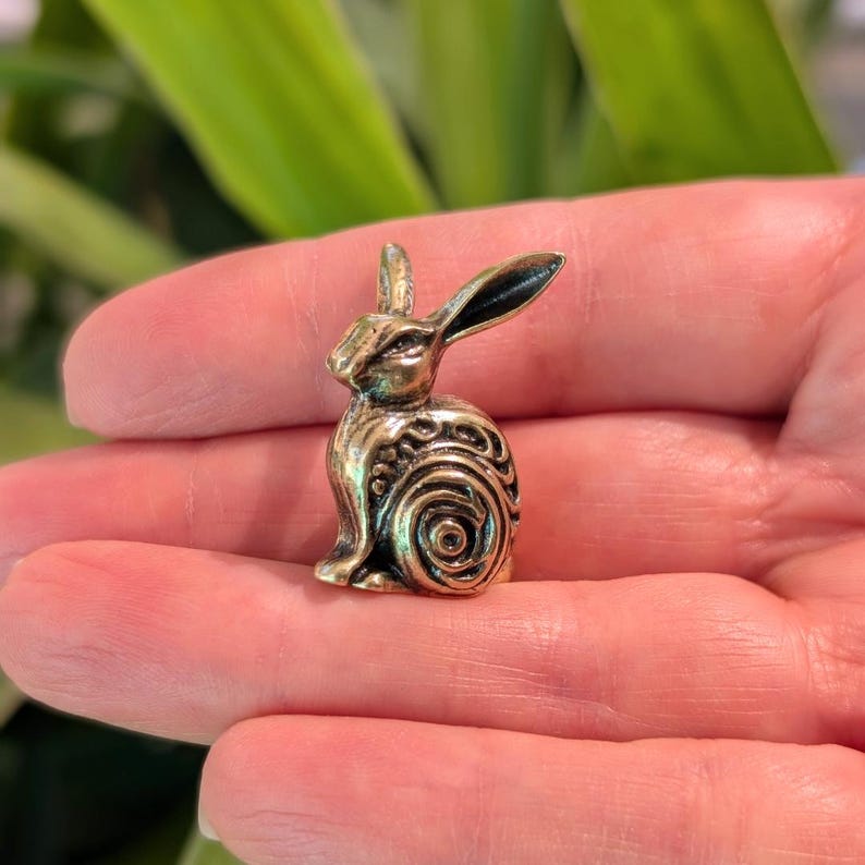 May include: A small, gold-colored metal rabbit figurine with intricate swirling patterns on its body. The rabbit is sitting upright with its ears perked up.