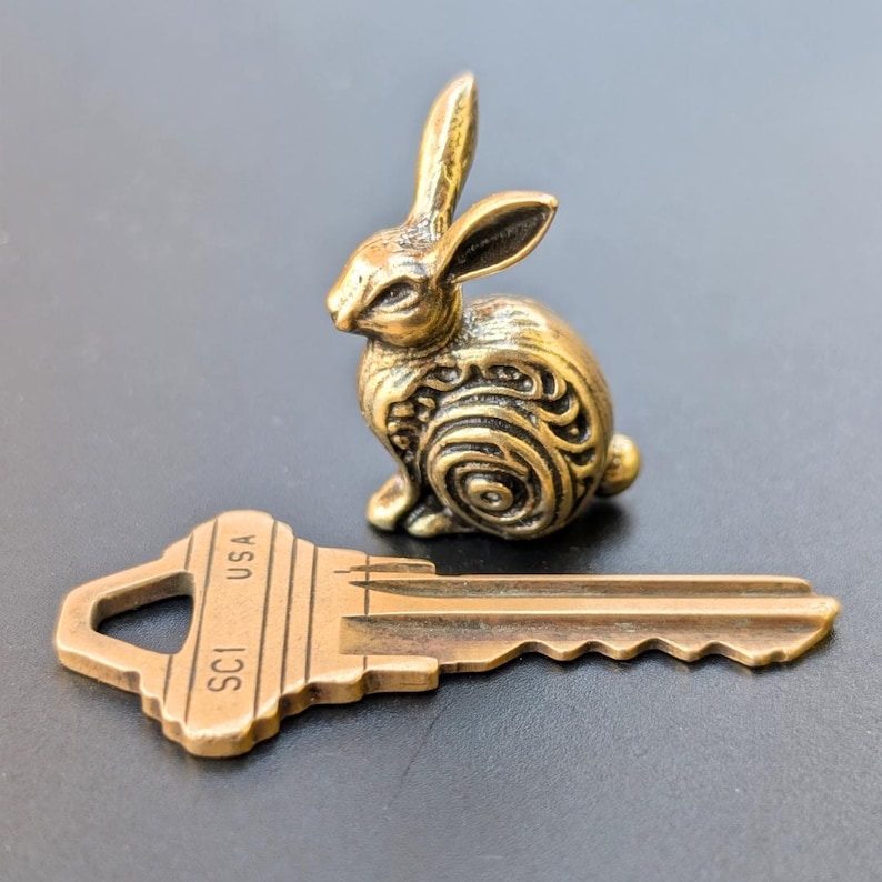 May include: A small, gold-colored, detailed rabbit figurine sits on a gray surface next to a brass key with the inscription "SCI USA".