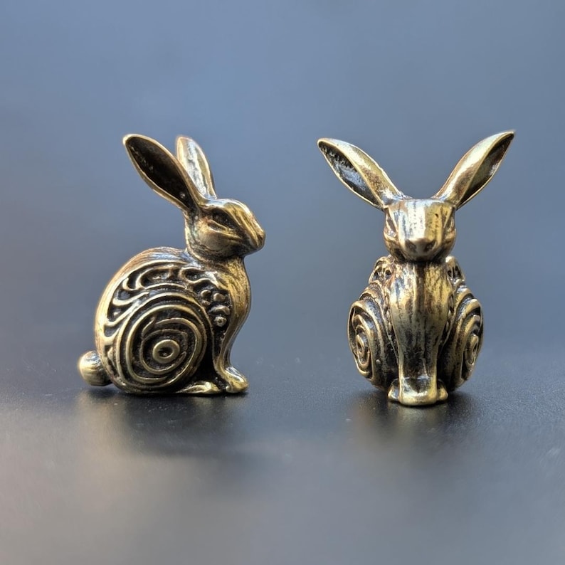 May include: Two small, gold-colored, metal rabbit figurines. The rabbits are sitting upright with their ears perked up. The rabbits have intricate, swirling patterns on their bodies.