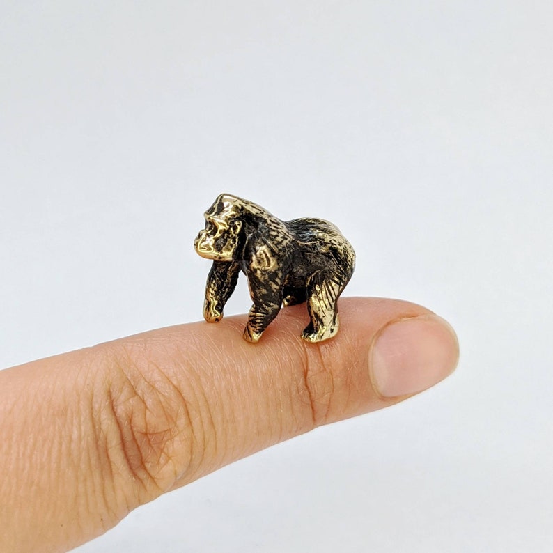 K&ouml;nnte beinhalten: Eine kleine, goldfarbene Figur eines Gorillas, der auf einem Finger steht. Der Gorilla ist schwarz und hat ein detailliertes Gesicht.