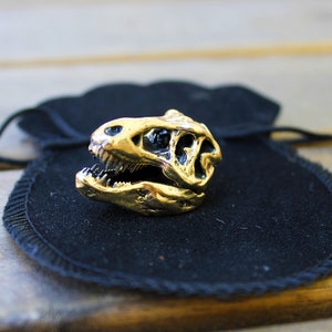 May include: A gold-colored dinosaur skull ring resting on a black velvet pouch.