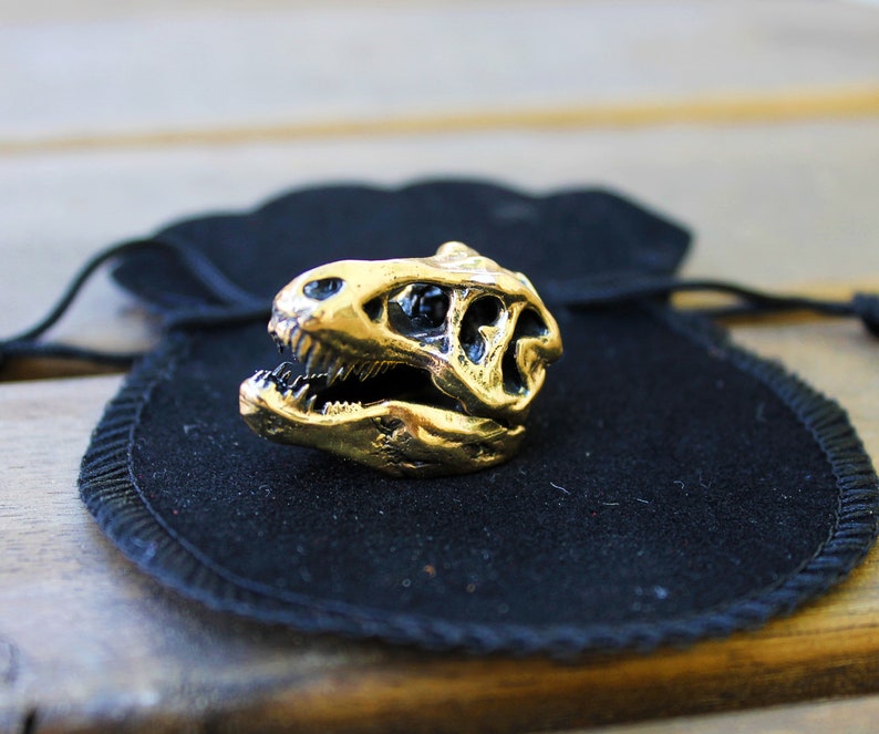 May include: A gold-colored dinosaur skull ring resting on a black velvet pouch.