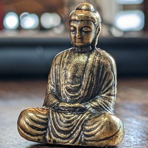 May include: A small, gold-colored statue of a meditating Buddha. The statue is sitting in a lotus position with its hands folded in its lap.
