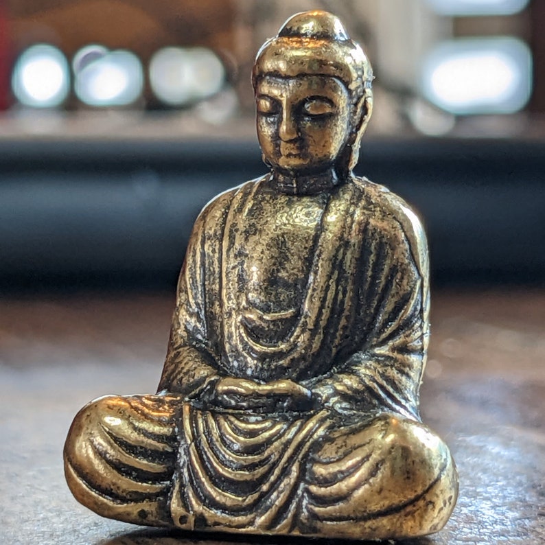 May include: A small, gold-colored statue of a meditating Buddha. The statue is sitting in a lotus position with its hands folded in its lap.