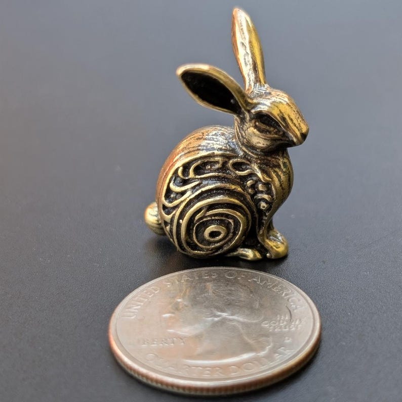 May include: A small, gold-colored metal rabbit figurine with intricate swirling patterns on its body. The rabbit is sitting upright with its ears perked up.