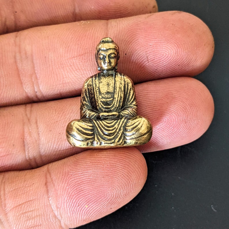 May include: A small gold-colored metal statue of a seated Buddha. The statue is detailed with intricate lines and folds in the clothing.