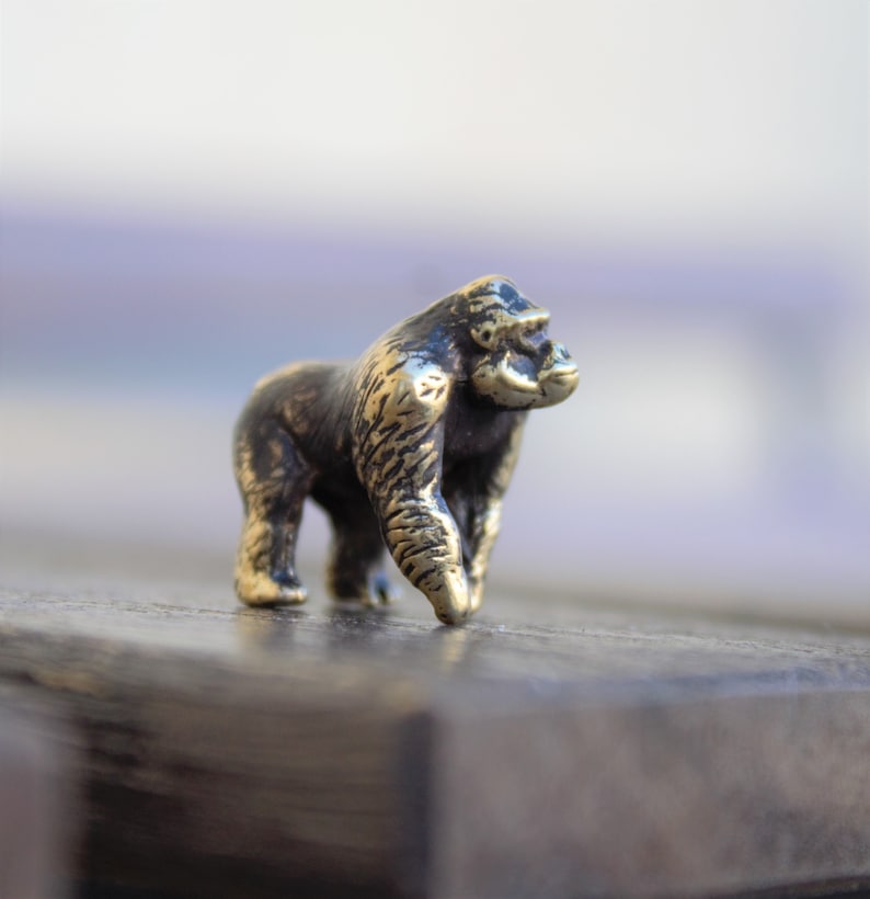 K&ouml;nnte beinhalten: Eine kleine, bronzefarbene Gorilla-Figur. Der Gorilla steht auf einer Holzoberfl&auml;che.