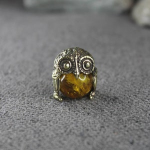 May include: A small brass owl figurine with an amber gemstone in the center. The owl has detailed wings and a round body.