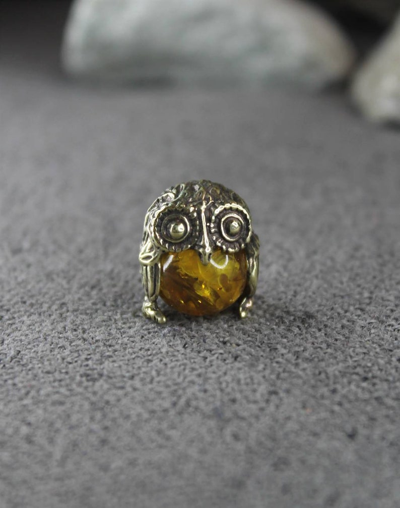 May include: A small brass owl figurine with an amber gemstone in the center. The owl has detailed wings and a round body.