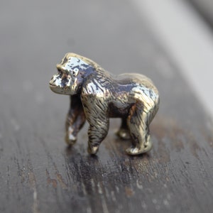 K&ouml;nnte beinhalten: Eine kleine, detaillierte, goldfarbene Metallfigur eines Gorillas, die auf einer Holzoberfl&auml;che steht.