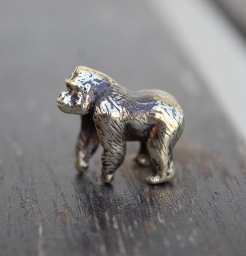 K&ouml;nnte beinhalten: Eine kleine, detaillierte, goldfarbene Metallfigur eines Gorillas, die auf einer Holzoberfl&auml;che steht.