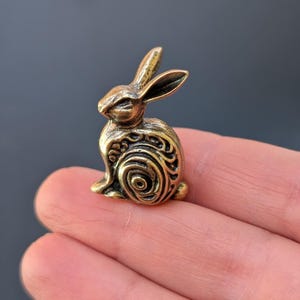 May include: A small, gold-colored metal rabbit figurine with intricate, swirling patterns on its body. The rabbit is sitting upright with its ears perked up.