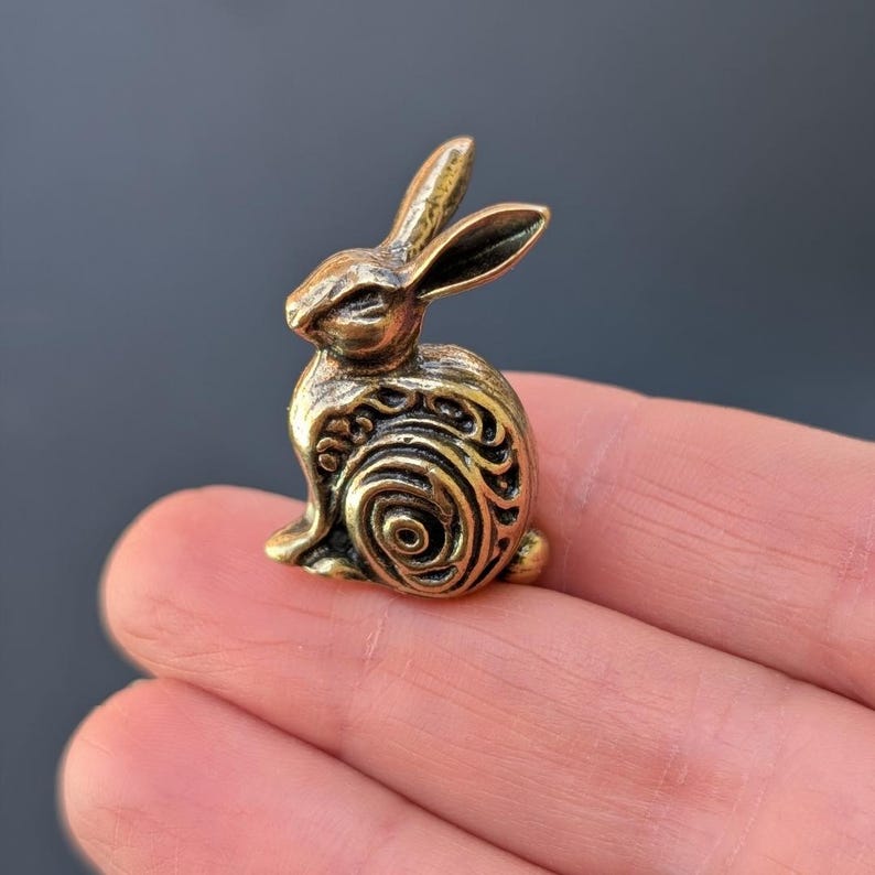 May include: A small, gold-colored metal rabbit figurine with intricate, swirling patterns on its body. The rabbit is sitting upright with its ears perked up.