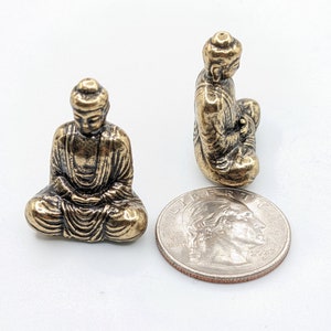 May include: Two small gold-colored metal statues of Buddha in a seated meditation pose. The statues are about the size of a US quarter.