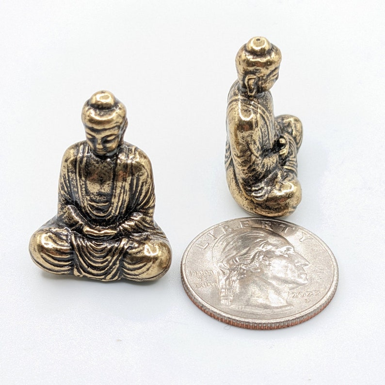 May include: Two small gold-colored metal statues of Buddha in a seated meditation pose. The statues are about the size of a US quarter.