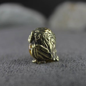 May include: A small, gold-colored owl figurine with intricate details. The owl is sitting on a gray textured surface.