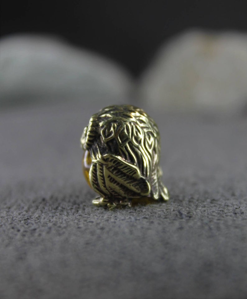 May include: A small, gold-colored owl figurine with intricate details. The owl is sitting on a gray textured surface.