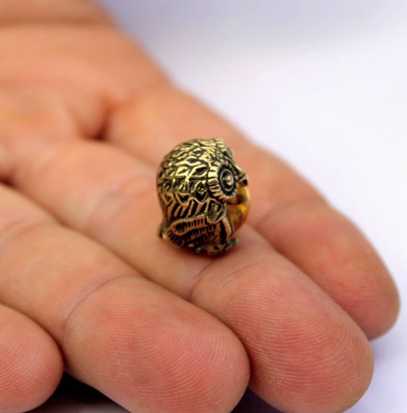 May include: A small, gold-colored owl figurine with intricate details. The owl is perched on a finger, showcasing its size.