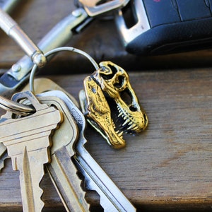 May include: A gold-colored dinosaur skull keychain attached to a silver key ring with multiple keys. The keychain is on a wooden surface.