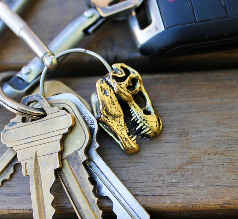 May include: A gold-colored dinosaur skull keychain attached to a silver key ring with multiple keys. The keychain is on a wooden surface.