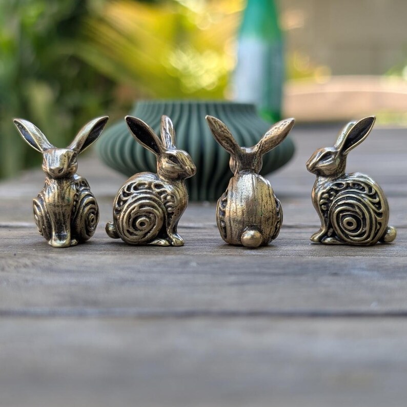 May include: Four small, detailed bronze rabbit figurines. Each rabbit is posed differently, with one facing forward, one facing backward, and two facing to the side. The rabbits have intricate designs on their bodies, including swirls and patterns.