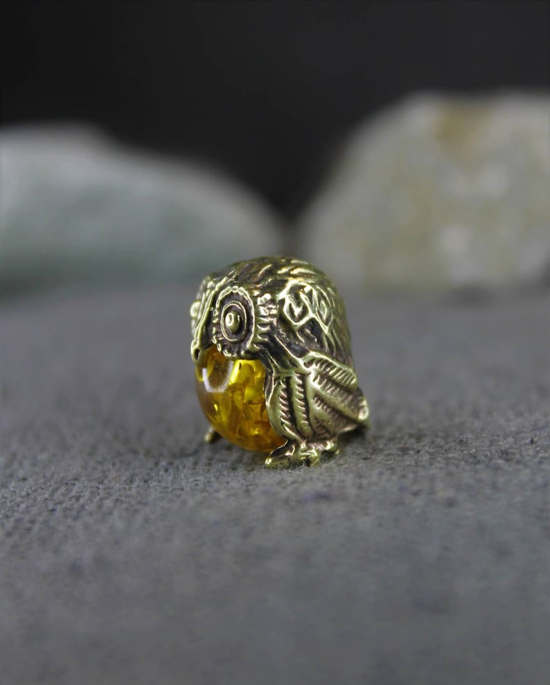 May include: A small, gold-colored owl figurine with a detailed design. The owl's eyes are made of amber.