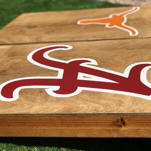 May include: Wooden cornhole boards with a light brown finish. One board features a large crimson "A" logo with a white outline, and the other board has an orange Longhorn logo. The boards are set on green grass.
