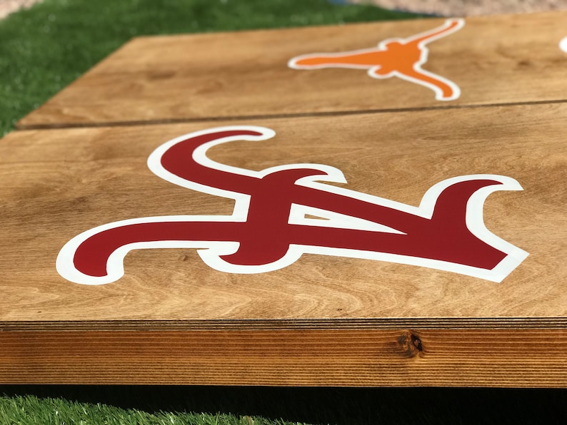 May include: Wooden cornhole boards with a light brown finish. One board features a large crimson "A" logo with a white outline, and the other board has an orange Longhorn logo. The boards are set on green grass.