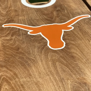 May include: A wooden cornhole board with a brown finish. The board features a white-rimmed hole and a large orange Longhorn logo with a white outline. The wood grain is visible.