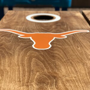 May include: Cornhole board with a natural wood grain finish. The board features a large orange and white Longhorn logo and a white-rimmed hole. The board is designed for outdoor games and activities.