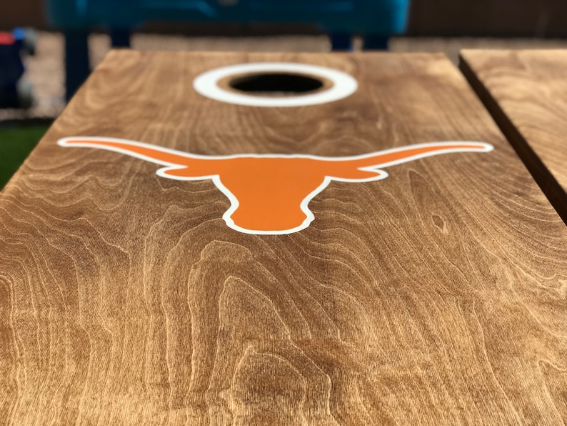 May include: Cornhole board with a natural wood grain finish. The board features a large orange and white Longhorn logo and a white-rimmed hole. The board is designed for outdoor games and activities.